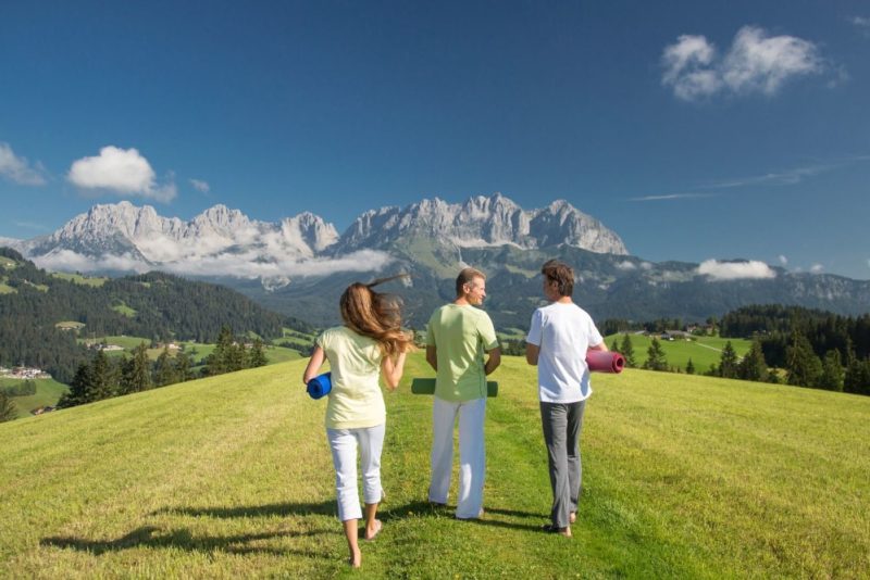 Drei Menschen in Yoga-Kleidung laufen auf einer grünen Wiese mit dem Wilden Kaiser im Hintergrund